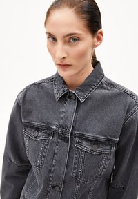 Eine Frau mit heller Haut und blauen Augen trägt eine verblasste schwarze Jeansjacke mit geknöpften Brusttaschen und blickt direkt nach vorne vor einem weißen Hintergrund.