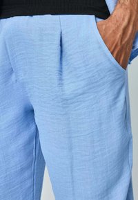 Pantalons bleu clair en tissu texturé, dotés de poches latérales et d'une coupe décontractée. Les détails de plis visibles ajoutent de la profondeur au design.