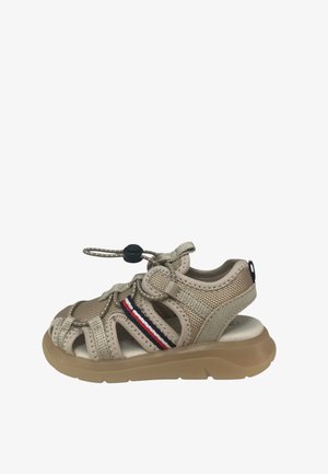 Παιδικό σανδάλι χρώματος μπεζ με κλειστή μύτη, αναπνεύσιμο δίχτυ, ρυθμιζόμενα ελαστικά κορδόνια και λεπτομέρεια με ρίγες κόκκινου, λευκού και μπλε στο πλάι της χοντρής σόλας.