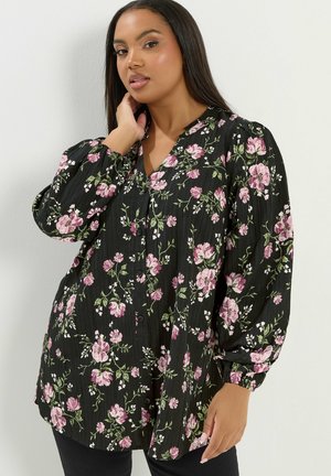 Kobieta z długimi ciemnymi włosami, ubrana w czarną kwiecistą bluzkę na guziki z długimi bufkami, pozująca z jedną ręką przy szyi.