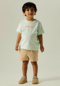 T-shirt à manches courtes vert menthe avec "OCEAN BLISS" en orange, associé à un short rayé orange et crème et des chaussures à enfiler en daim gris.