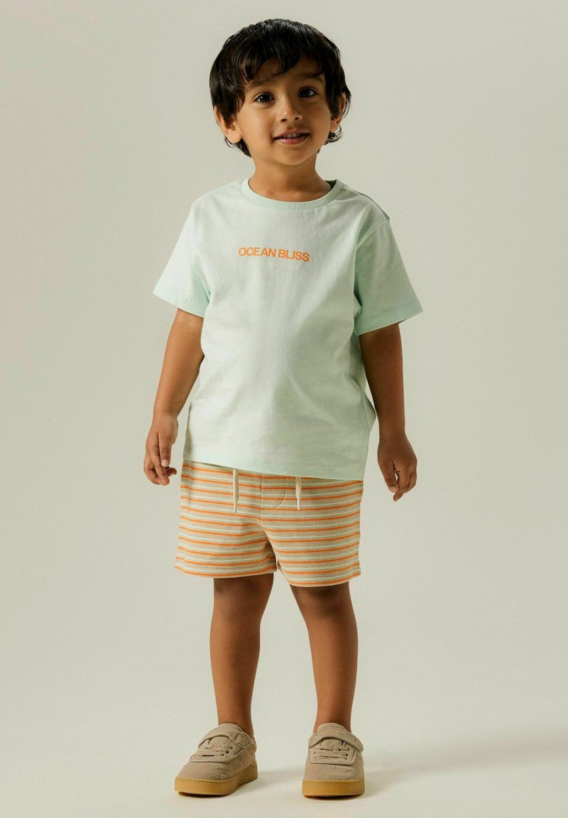 T-shirt à manches courtes vert menthe avec "OCEAN BLISS" en orange, associé à un short rayé orange et crème et des chaussures à enfiler en daim gris.