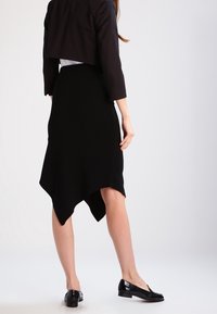 Jupe noire en tricot avec un ourlet asymétrique, associée à une veste noire courte. Le mannequin porte des mocassins noirs, affichant une silhouette élancée.