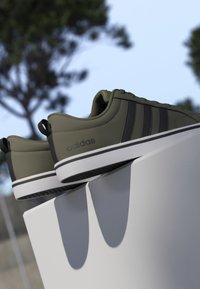 Par de zapatillas Adidas de color verde oliva con rayas negras descansando sobre un saliente blanco al aire libre con árboles al fondo.