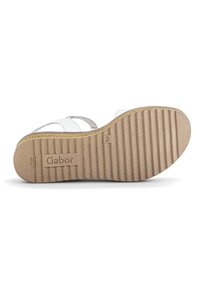 White sandal with a textured sole, featuring soft straps and a rubberized midsole. The bottom has a ribbed pattern and logo detail.
