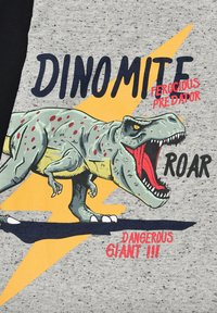 Graues T-Shirt mit illustriertem T-Rex-Dinosaurier, roten Flecken, offenem Mund und dem Text "DINOMITE ROAR DANGEROUS GIANT", hervorgehoben durch einen gelben Blitz.