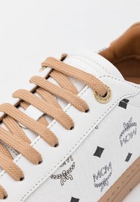 Baskets en cuir blanc avec surface texturée, lacets marron clair et détail logo doré. Présente des motifs noirs imprimés incluant des diamants et du marquage.