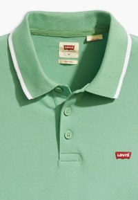Polo vert avec un tricot texturé, deux boutons et un col rayé blanc. Présente un logo rouge Levi's sur la poitrine gauche. Taille M.