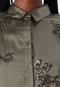 Camisa de satén verde oliva con cierre de botones al frente, cuello con solapa y bordado floral en lentejuelas negras en el lado izquierdo.