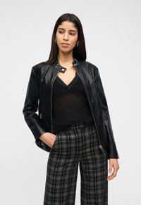 Γυναίκα με μακριά σκούρα μαλλιά που φοράει μαύρο υφασμάτινο δερμάτινο μπουφάν, μαύρη διάφανη μπλούζα με V-λαιμόκοψη και ασπρόμαυρο καρό παντελόνι, με το χέρι στην τσέπη.