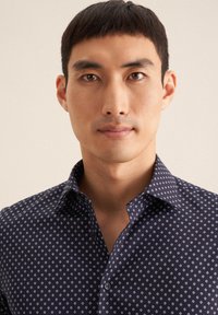 Donkerblauwe shirt gemaakt van stof met een ruitpatroon in lichtblauw. Heeft een puntige kraag en een knoopsluiting aan de voorkant.