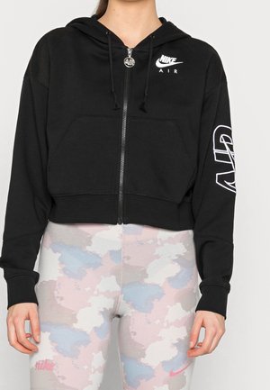 Osoba ubrana w czarną bluzę Nike Air na zamek z kapturem oraz jasne pastelowe legginsy Nike z kamuflażowym wzorem, stoi z rękami swobodnie opuszczonymi wzdłuż ciała.