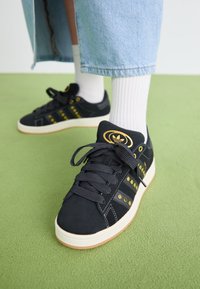 Melni zamšas sporta apavi ar zelta akcentiem, aprīkoti ar apaļu logotipu, biezām auklām un gumijas zoli. Valkāti ar baltām sporta zeķēm un džinsiem.