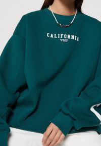 Sweat-shirt bleu sarcelle avec poignets côtelés et encolure ajustée, arborant le texte blanc "CALIFORNIA" et des détails de marque. Accessoirisé d'un collier de perles.