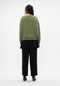 Sudadera verde con hombros caídos, combinada con una falda negra plisada y pantalones negros de pierna ancha. Textura suave y corte relajado.