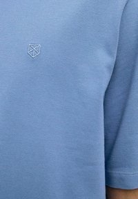 Šviesiai mėlyna polo marškinėliai su tekstūruotu mezgimo audiniu ir mažuoju išsiuvinėtu logotipu ant krūtinės. Trumpomis rankovėmis ir klasikinio apykaklės dizainu.