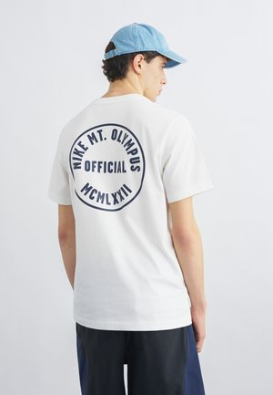 Νέος άνδρας που φοράει λευκό T-shirt Nike με μαύρο λογότυπο του Όρους Όλυμπος και ρωμαϊκούς αριθμούς στην πλάτη, μπλε καπέλο και σκούρα σορτς.