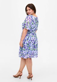 Blumenkleid in hellem Lavendel mit grünen Akzenten; hat kurze Ärmel, eine taillierte Taille und einen fließenden Rock mit lila Blumen.