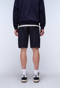 Man die zwarte cargoshorts, witte sokken en zwart-witte sneakers draagt, van achteren gezien tegen een effen lichtgrijze achtergrond.