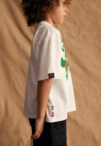 Weißes Baumwoll-T-Shirt mit grünem Grafikdesign und braunen Akzenten. Kurze Ärmel, lockere Passform. Schwarze Cargo-Shorts, rotes Logo am Saum.