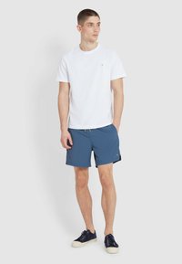 Weißes Baumwoll-T-Shirt, blaue Badeshorts mit elastischem Bund und Kordelzug sowie marineblaue Turnschuhe mit weißen Sohlen, vor einem schlichten Hintergrund stehend.