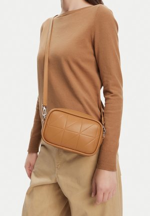 Mujer con suéter de manga larga color marrón claro y pantalones beige, llevando un bolso cruzado acolchado de cuero marrón claro con una correa ajustable.