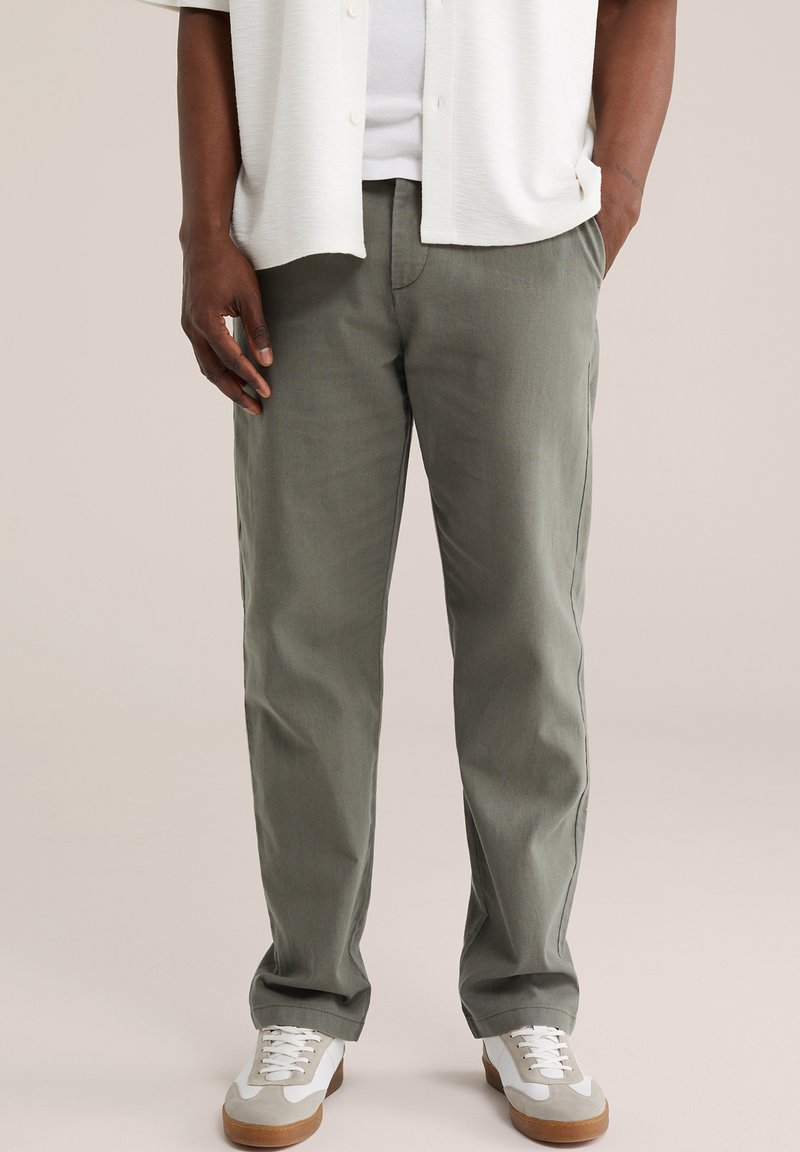 Mees, kes kannab halli sirge lõikega pükse, valgeid tossusid ja valget särk, üks käsi taskus ja teine lõdvestunult külje kõrval.