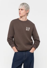Brown sweatshirt with long sleeves and crew neck, featuring a white "GAP" logo on the chest. Paired with dark blue jeans.