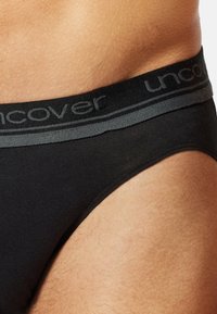 Schwarze Unterhose aus weichem Stoff, mit einem grauen elastischen Bund, der das Logo "uncover" trägt. Glatte Textur mit minimalistischem Design.