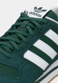 Grøn Adidas-sneaker med hvide striber og matchende grønne snørebånd, der viser tekstureret stof og ruskindsdetaljer på oversiden.