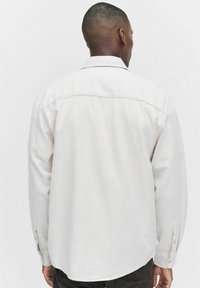 Lys beige langærmet skjorte med knapmanchetter, blødt stof, standard krave og en let buet kant, vist fra ryggen.