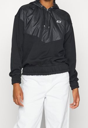 Personne portant un sweat à capuche noir avec un panneau brillant sur la poitrine et un pantalon blanc, mains détendues le long du corps.
