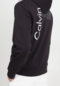 Zwarte hoodie met een voorzak in kangoeroestijl. Groot wit logo verticaal op de achterkant gedrukt. Katoenmix stof. Klassieke pasvorm met geribde manchetten.