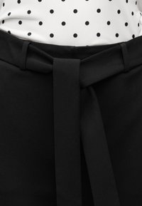 Μαύρο παντελόνι ψηλόμεσο με φαρδιά, δεμένη ζώνη, φορεμένο πάνω από ένα λευκό πουκάμισο με μαύρες πουά βούλες.