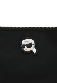 Pochette en nylon noir avec un graphisme en relief d'une silhouette aux cheveux blancs et portant des lunettes de soleil, fermée par une fermeture éclair argentée.