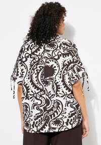 Blouse à manches courtes avec un motif paisley noir et blanc, présentant des accents à nouer sur les manches et une fermeture à bouton au dos. Tissu léger.
