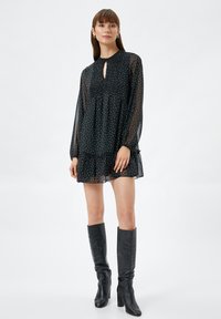 Schwarzes Kleid mit weißen Punkten, langen transparenten Ärmeln, gerafftem Oberteil, Schlüsselloch-Ausschnitt und Rüschensaum, getragen mit schwarzen kniehohen Stiefeln.