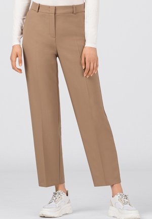 Femme portant un pantalon beige droit avec un pli sur le devant et des baskets blanches épaisses, debout devant un fond clair uni.