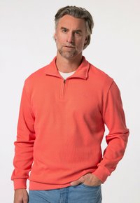 Korálový pulovr s polo zipem, žebrovanou texturou, dlouhými rukávy a malým logem na hrudi. Nosený s světle modrými džíny.