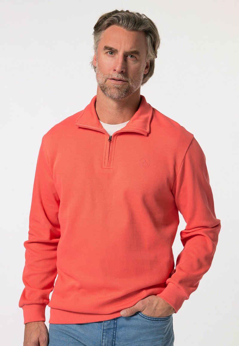 Korálový pulovr s polo zipem, žebrovanou texturou, dlouhými rukávy a malým logem na hrudi. Nosený s světle modrými džíny.