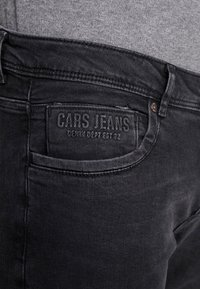 Jeans de mezclilla negra que presentan una gran etiqueta en relieve en el bolsillo trasero que dice "CARS JEANS", con costuras en contraste y una textura suave.