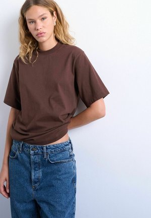 Jeune personne aux cheveux blonds ondulés portant un t-shirt marron et un jean en denim bleu, debout contre un mur blanc.