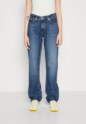 Osoba ubrana w luźne, niebieskie dżinsy, biały top na ramiączkach, białe trampki z żółtymi akcentami, stojąca na tle jasnego, jednolitego tła.