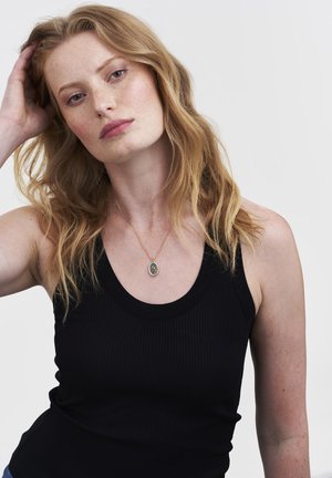 Crni prsluk s rebrastim uzorkom i okruglim izrezom na vratu. Tanka zlatna ogrlica s ovalnim privjeskom ukrašenim šarenim detaljima.