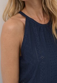 Kvinna med långt blont hår som bär en ärmlös klänning i marinblå spets som visar skuldra och del av halsen mot en neutral bakgrund.