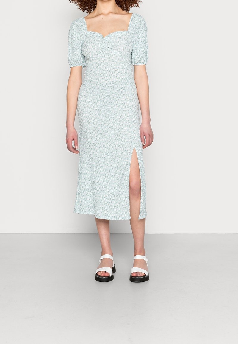 Femme portant une robe midi bleu clair avec des manches courtes bouffantes, un motif floral, une fente à l'avant, et des sandales blanches avec des semelles noires.