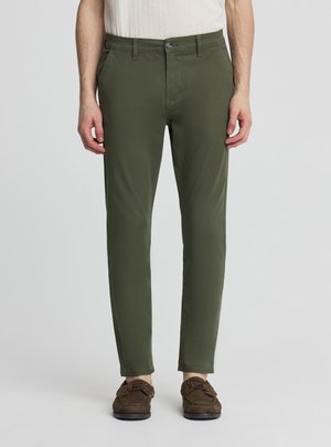 Uomo che indossa pantaloni slim fit verde oliva e mocassini marroni, in piedi davanti a uno sfondo chiaro e semplice.