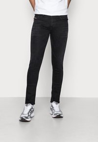 Svarta slim-fit jeans med en lätt glans, femfickorsdesign och subtila sömdetaljer, i kombination med gråa sportskor.