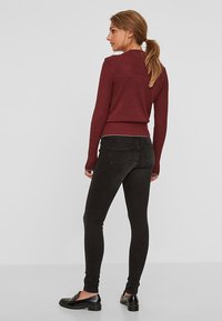 Suéter de canalé burdeos, diseño ajustado, combinado con jeans skinny negros. Zapatos planos negros. Fondo liso, luz natural.