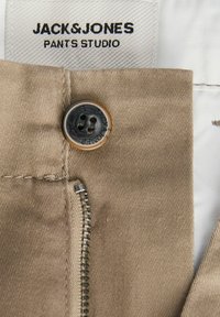 Beige παντελόνι με κουμπί και φερμουάρ, φέρει ετικέτα "JACK & JONES PANTS STUDIO" στο εσωτερικό της ζώνης.